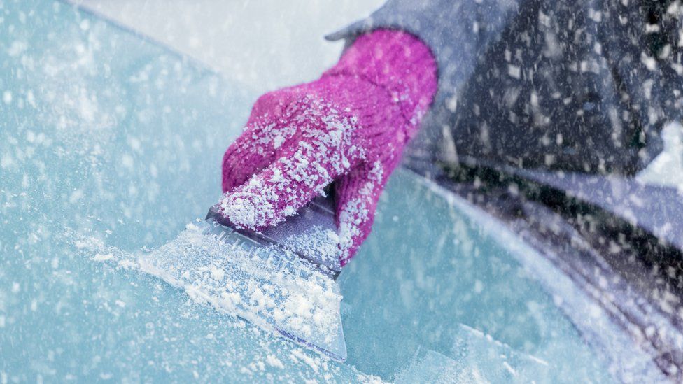 A woman wearing a pink glove scrapes ice off a windscreen